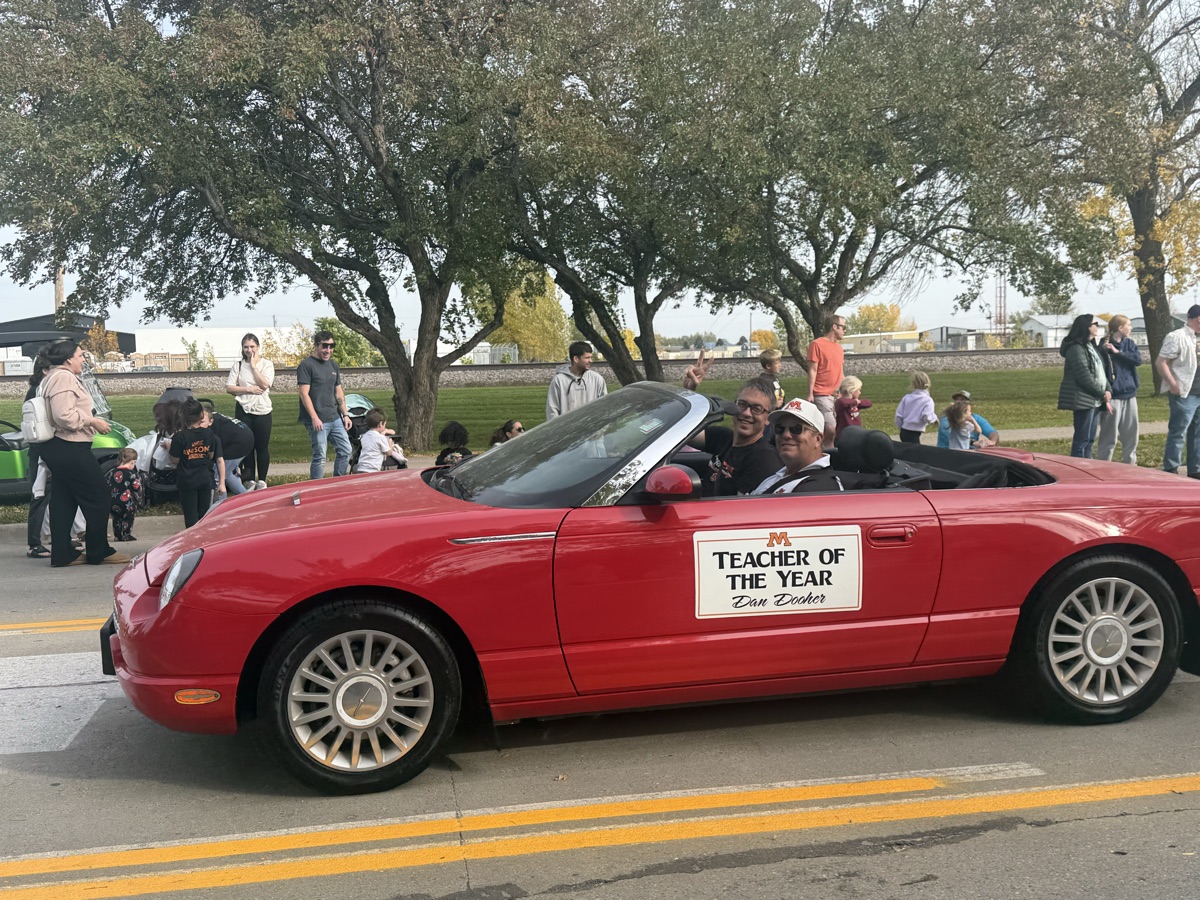 Dan Dooher riding in a red convertible in the homecoming parade behind a Teacher of the Year placard
